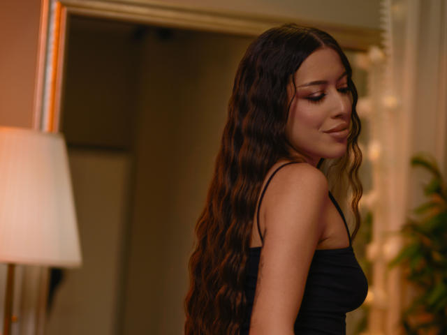 AlaraMyst with long wavy brown hair, wearing a black spaghetti-strap top, looking down with a subtle smile, soft lighting, warm indoor setting, visible lamp on left.