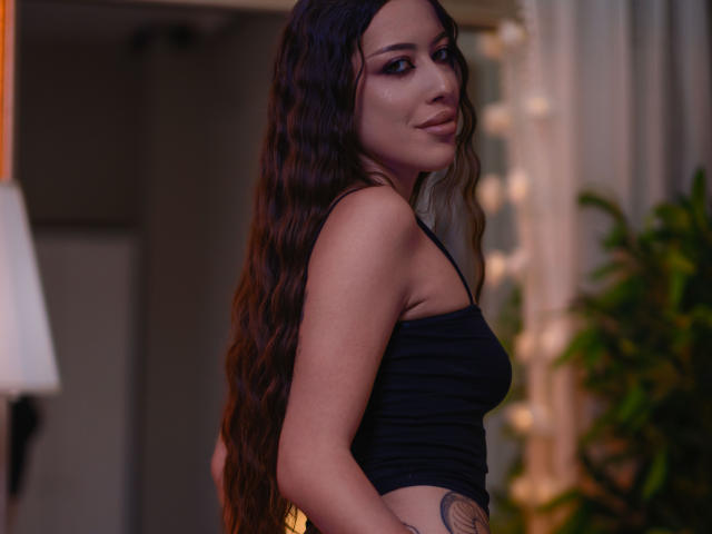 AlaraMyst, avec de longs cheveux bruns ondulés, portant un crop top noir, montrant un tatouage sur son côté droit, regardant par-dessus son épaule, avec un léger sourire..