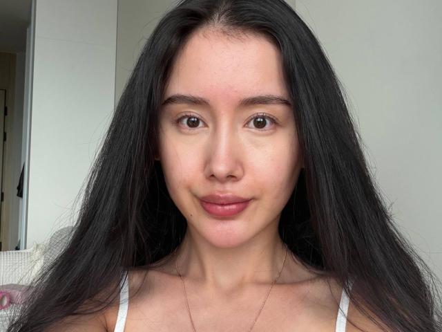 Kandace Moles hat langes, schwarzes Haar, braune Augen und ein leichtes Lächeln. Sie trägt ein weißes Oberteil mit kleinen Brüsten und eine dünne Halskette. Ihre Haut ist hell..