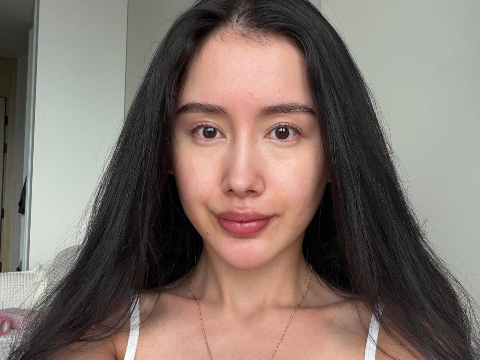 KandaceMoles heeft lang zwart haar, bruine ogen en een lichte glimlach. Ze draagt een witte top met een kleine decolleté en een dunne ketting. Haar huid is licht..