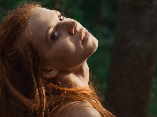 FoxyyMolly, con i capelli rossi, lentiggini e pelle chiara, guarda verso l'alto con un leggero sorriso, mostrando il collo e la parte superiore del petto. Il suo seno è parzialmente visibile, all'aperto..