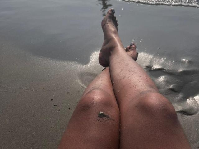 EsmeraldaRoe's sandy, sun-kissed legs stretched out on the beach, feet touching gentle waves, with small patches of sand sticking to her skin.