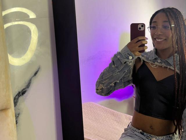 EsmeraldaRoe with long braids, glasses, black crop top, and denim jacket, holding phone in mirror selfie, smiling, showing her toned midriff and small tits.