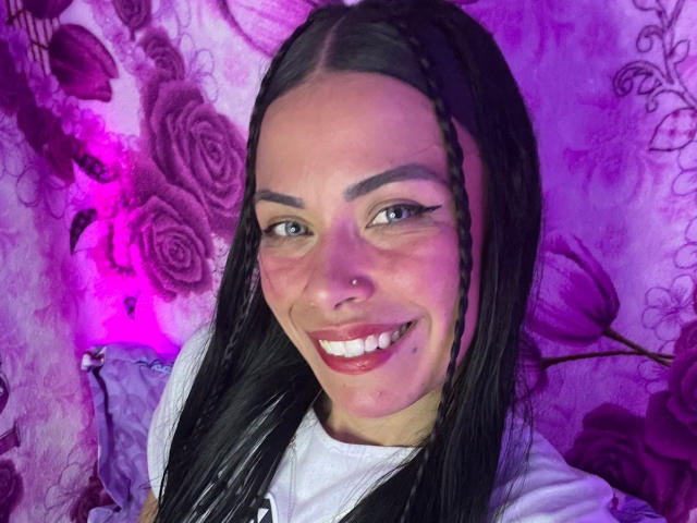 ZoeLux, con trenzas negras largas, ojos azules y un piercing en la nariz, sonriendo con dientes blancos y brillantes, vestida con una camisa blanca, y un fondo con un estampado de rosas moradas..