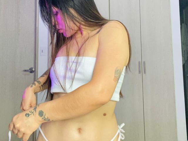 SophiaMarx with long brown hair, white strapless top, white bikini bottoms, visible tattoos on arms, looking down, pink light on face, wooden door in background, holding white string.