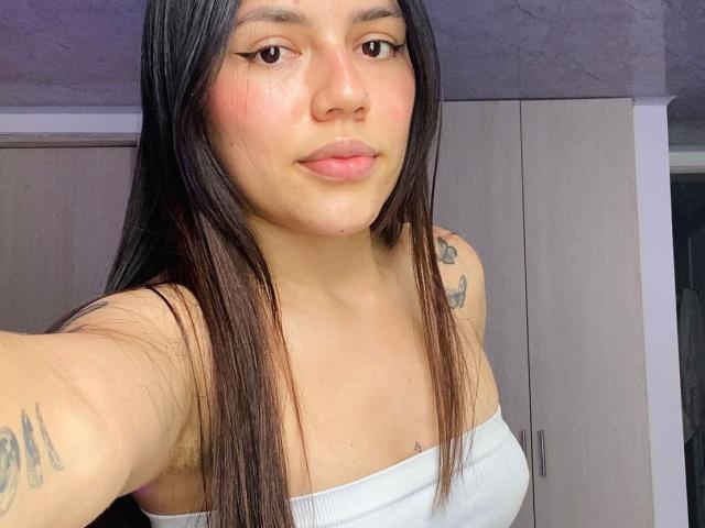 SophiaMarx has straight black hair, light brown skin, small tattoos on arms, wearing a white strapless top, showing her tits, with a subtle smile and pink blush.
