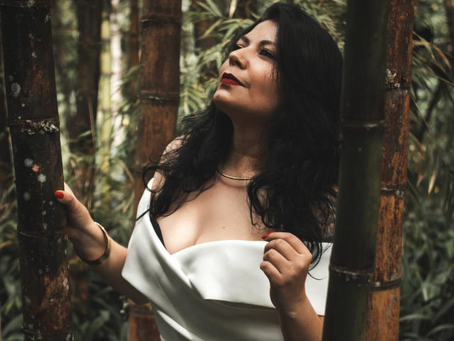 AnnaKerly, con cabello negro, lápiz labial rojo y vestido blanco, sosteniendo bambú, mostrando sus senos, mirando hacia arriba, con las manos en el vestido, fondo de bosque, segura de sí misma, sexy, plano medio..