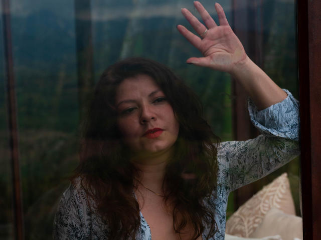 Annas Hand liegt auf einem Glas, ihr welliges, braunes Haar und der rote Lippenstift bilden einen Kontrast zu ihrer Spitzenbluse, die ihre weichen Brüste und ihre dezenten Dekolletés zeigt..