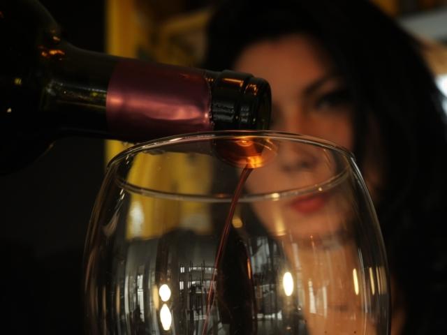 AnnaKerly está servindo vinho tinto de uma garrafa escura em um copo transparente, com o rosto levemente desfocado ao fundo, cabelos escuros e batom vermelho..