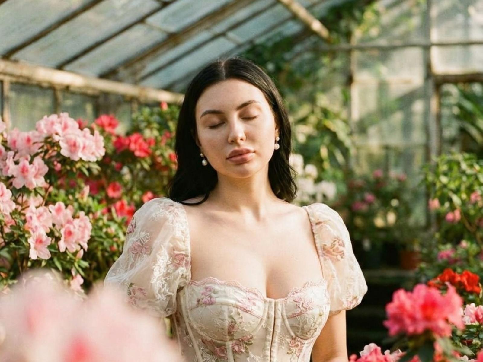 Judy Goyal, mit geschlossenen Augen, trägt ein blumiges Spitzenkleid und zeigt ihre Brüste. Sie ist von rosa und roten Blumen in einem gläsernen Gewächshaus umgeben. Sie hat dunkles Haar und Perlenohrringe..