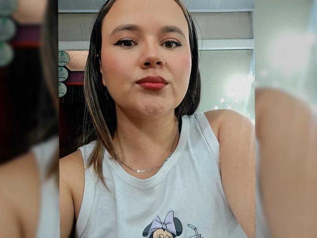NatalieRey has puffy red lips, a nose ring, and wears a white tank top with a Minnie Mouse print. She's in front of a mirror, looking cute.