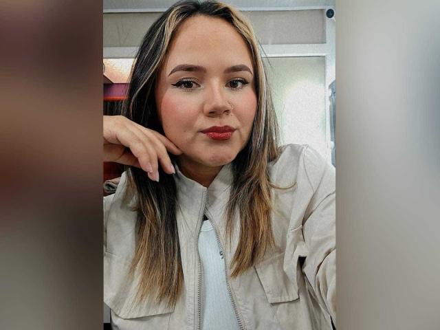 Natalie Rey, com cabelos castanho-claros com mechas loiras, vestindo uma jaqueta bege sobre uma camisa branca, usa batom vermelho e tem um leve semblante franzido, com o queixo apoiado na mão..