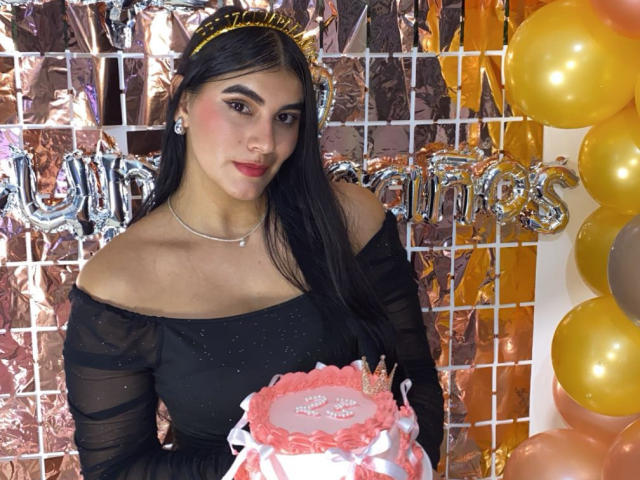 AlaraKey has long black hair, a gold headband, black off-shoulder dress, holding pink cake with white ribbon, wearing silver necklace, surrounded by gold balloons.