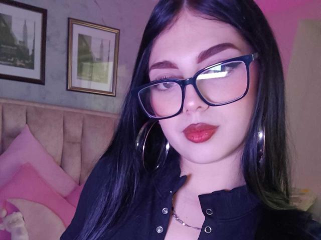 Isabella Rossi, com cabelo preto, óculos, batom vermelho, brincos de argola, camisa preta, abotoada, pele clara, olhando para a câmera, em um quarto, com travesseiros rosa, e quadros na parede..