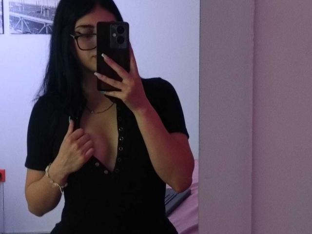 IsabellaaRossi taking a mirror selfie, wearing glasses, black shirt with buttons, exposing her tits, holding phone, long black hair, bracelet, necklace, in dimly lit room.