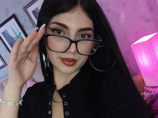 Isabella Rossi met lang zwart haar, die zwarte brillen draagt, een zwarte blouse, rode lippenstift en oorringen, en een glas vasthoudt met teal gekleurde nagellak, terwijl ze glimlacht. In de achtergrond staat een roze lamp..