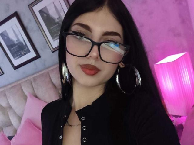 Isabella Rossi, avec des lunettes, du rouge à lèvres rouge, des cheveux noirs, de grandes boucles d'oreilles et un haut noir boutonné, laissant légèrement entrevoir ses seins. Une lampe rose est visible en arrière-plan..