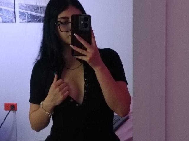 Isabella Rossi, met zwart haar en een bril, draagt een zwarte top met een diepe V-hals, houdt een telefoon voor haar gezicht en toont haar borsten in een spiegelzelfportret..