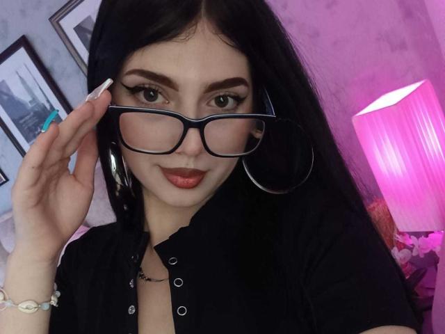 Isabella Rossi, avec ses cheveux noirs, ses lunettes et ses boucles d'oreilles, ajuste ses lunettes, porte une chemise noire et du rouge à lèvres, dans une pièce éclairée en rose, et affiche un sourire discret..
