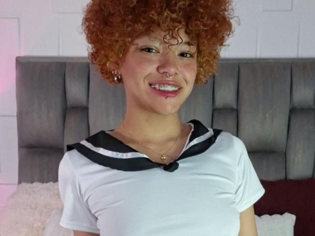 KeiryGinger, avec ses cheveux roux bouclés, souriante, portant une chemise blanche avec des détails noirs, un anneau au nez et un collier en or, se tient devant une tête de lit grise recouverte de tissu..