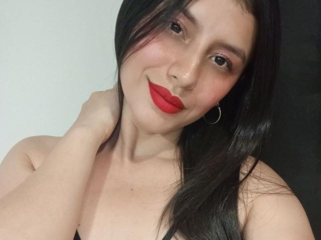 MadissonFontaine with red lipstick, black hair, hoop earrings, and a hand on her neck, smiling at the camera. Her tits are visible, showing a black strap top.