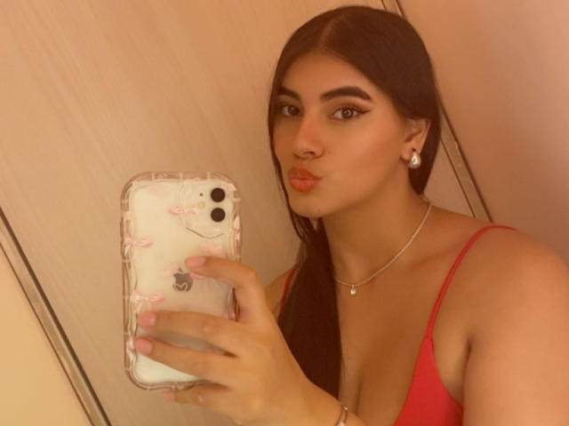 AlaraKey with pouty lips, dark brown hair, and red top, holding phone with pink nail polish, wearing pearl earrings, and necklace, in selfie with her ass visible.