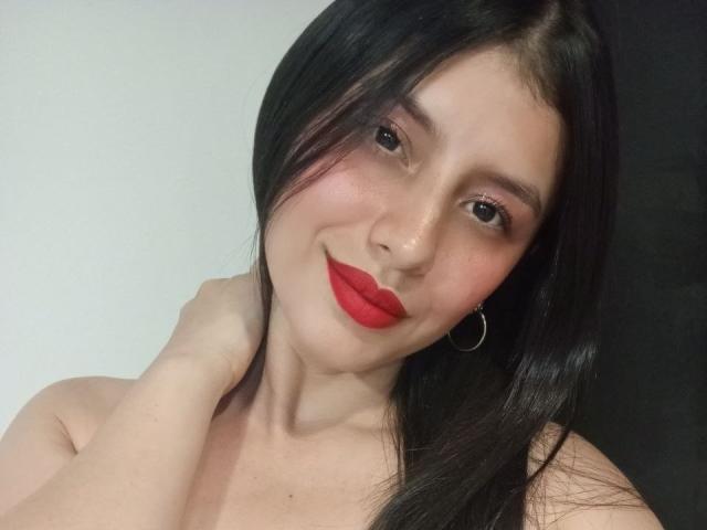MadissonFontaine with red lipstick, black hair, hoop earrings, and hand on neck, smiling at camera, showing bare shoulders, light skin, and cute face.