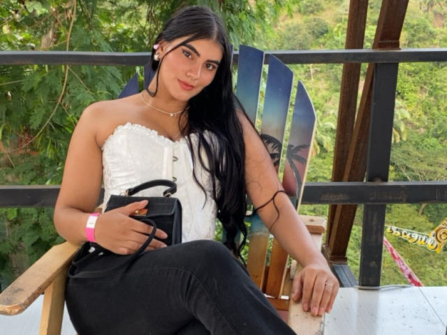 AlaraKey with long black hair, white strapless top, black pants, holding black camera, pink wristband, sitting on wooden chair, green leafy background, relaxed, cute, small ass.