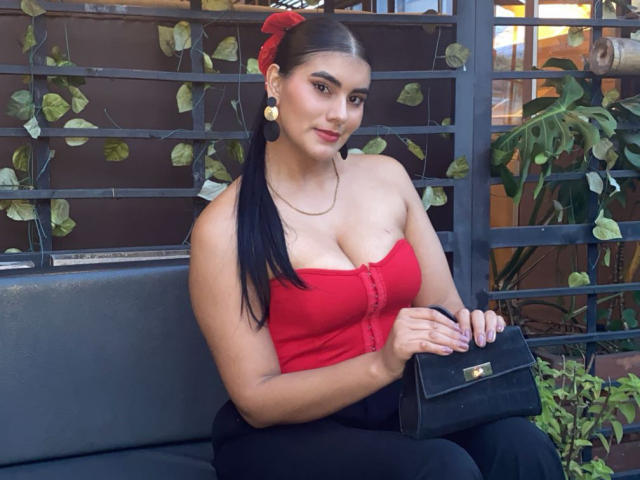 AlaraKey with long black hair in a ponytail, wearing a red strapless top, showing her tits, holding a black purse, sitting on a black bench, smiling.