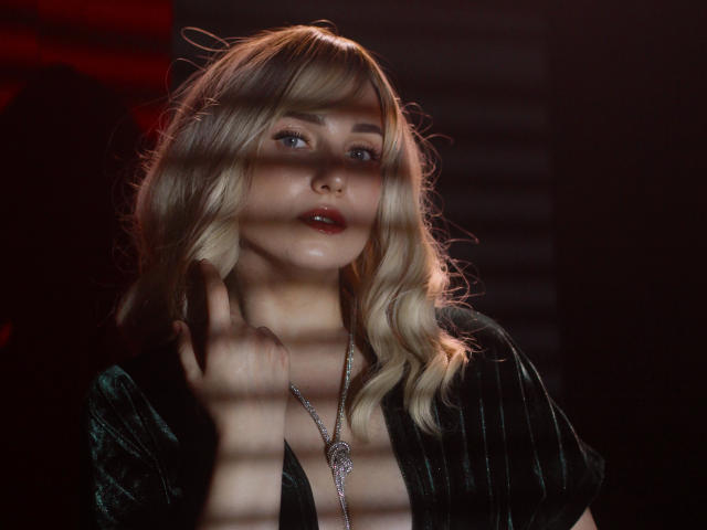 MilenaBaldwin with blonde, wavy hair, wearing a black, pinstriped dress, has a silver necklace, and her hand near her neck, with red light and shadow on her face.