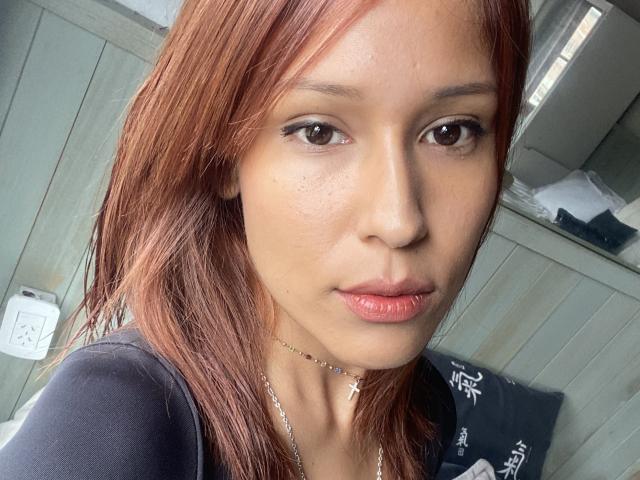 KiaraHernandezX has red hair, brown eyes, and is wearing a black shirt with a silver necklace. She has a small, cute nose and slightly parted pink lips.