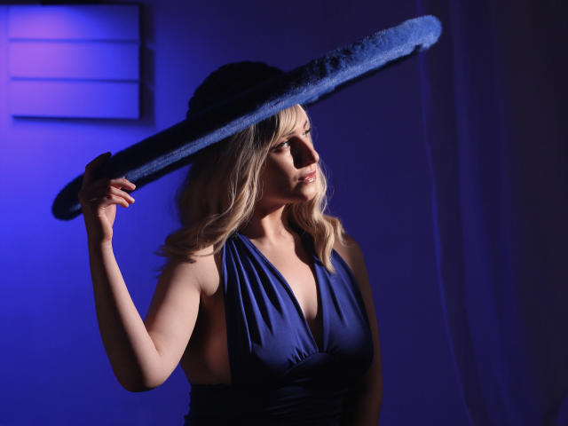 MilenaBaldwin with blonde hair, wearing a blue dress, holding a blue baseball bat over her shoulder, blue hat on head, in a dimly lit, blue-tinted room.