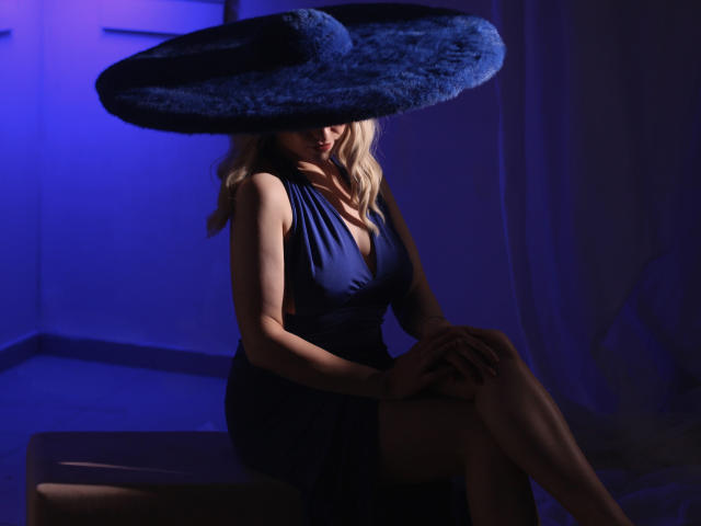 MilenaBaldwin with blonde hair, wearing a dark blue dress and huge blue fur hat, sitting in blue-lit room, legs crossed, hands on knees, showing her tits slightly.