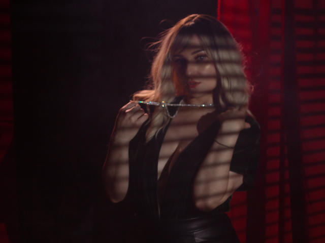 MilenaBaldwin, com cabelo loiro, segurando um colar, vestindo uma blusa preta com decote profundo em V, com sombras de persianas no rosto, cortina vermelha ao fundo, aparência escura, sexy e confiante..