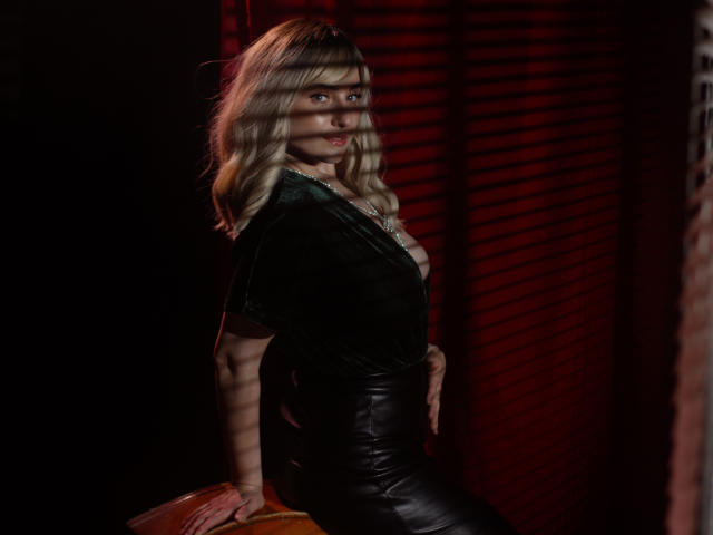 MilenaBaldwin with blonde hair, green velvet top, and black leather skirt, leaning on a yellow chair, red-striped shadows on her face, looking over her shoulder with a slight smile.