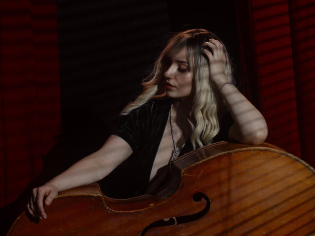 MilenaBaldwin with blonde hair, black dress, and hand in hair, leaning on a large wooden bass, striped light shadows, looking down, dark red curtains, sensual, sexy, tits visible.