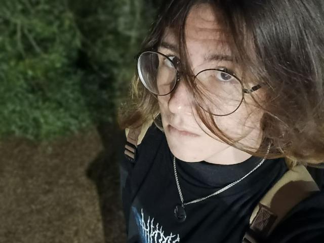 ReaRosine mit Brille, braune, wellige Haare, ernster Gesichtsausdruck, trägt ein schwarzes Hemd, einen Tarnrucksack, eine Halskette, hat helle Haut, im schwachen Außenlicht..