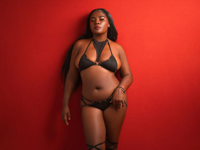 NylahGlow with dark skin and long black hair wearing a black strappy bikini, showing her big ass and small, round tits, standing against a red wall.