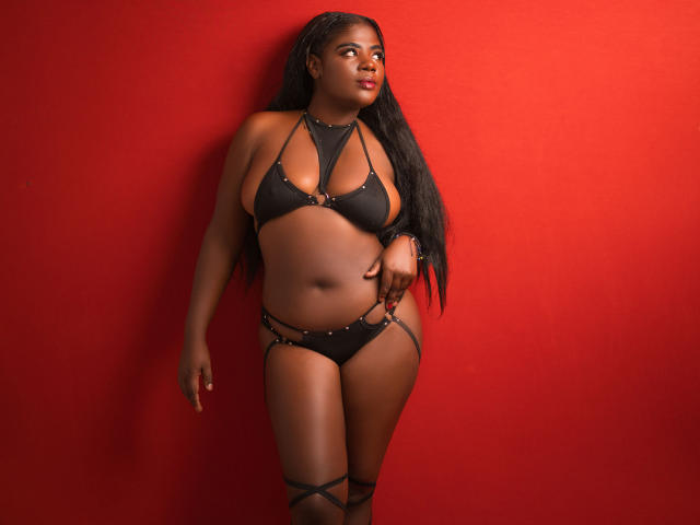 NylahGlow, met donkere huid en lang zwart haar, draagt een zwart bikini met bandjes, staat tegen een rode muur, kijkt naar de zijkant met een zelfverzekerde uitdrukking en laat haar grote achterwerk zien..
