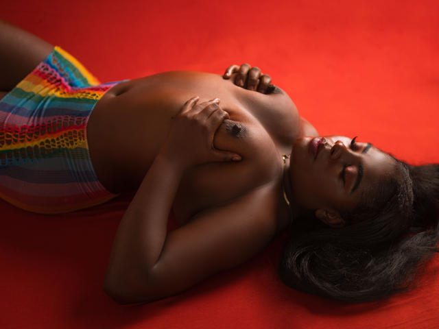 NylahGlow, oben ohne, liegt auf Rot, bedeckt ihre dunkelbraunen Brüste mit den Händen, bunte, gestreifte Hose, schwarzer Choker, dunkelbraune Haut, langes, schwarzes Haar, Augen geschlossen, entspannt, sexy..