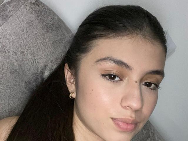 IsabelaRios has light brown skin, dark brown eyes, and long black hair. She's wearing small gold earrings, with a subtle pout and slightly upturned nose. Her tits are not visible.