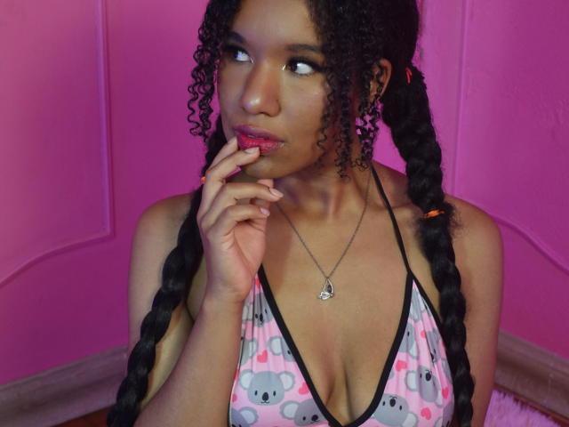 KatalinaGildas, avec des tresses noires bouclées, portant un haut de maillot de bain à motifs roses et gris, mordant son doigt, regardant sur le côté, peau foncée, collier avec un pendentif en forme de cœur, mur rose..