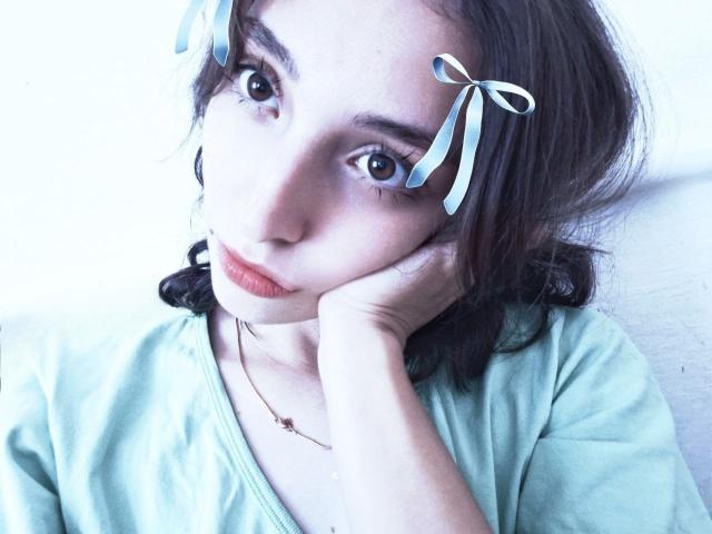 Leonie Rahim, avec sa peau pâle, ses yeux bleus et ses cheveux bruns ondulés, porte une chemise verte, a des rubans dans les cheveux, une main posée sur sa joue, et a l'air triste. Un éclairage bleu doux..