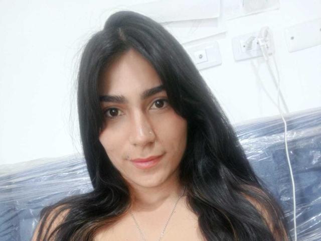 ZoeeRoux has long black hair, brown eyes, and a small nose ring. She's smiling with a slight blush, wearing a silver necklace, and has visible makeup on her face.