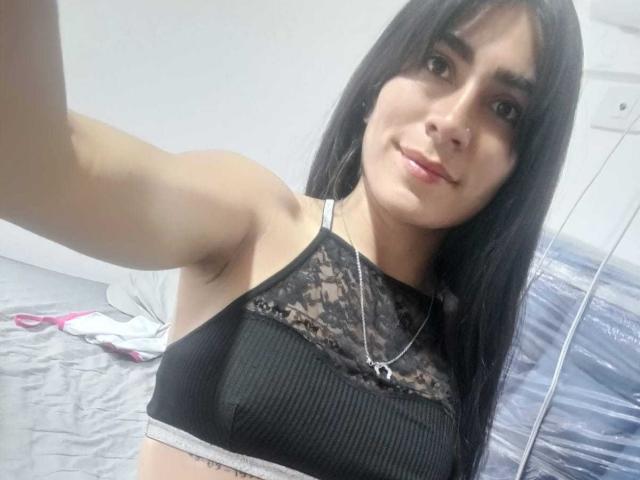 ZoeeRoux with long black hair, wearing a black lace bralette, smiling at the camera, showing her small tits, and a silver necklace in a selfie.