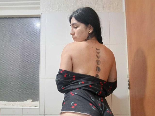 ZoeeRoux with black hair, back tattoo of five moon phases, and cherry-patterned black top, exposing her bare back and ass. She's looking over her shoulder, smiling.