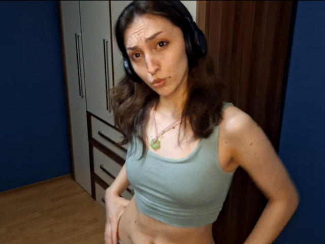 LeonieRahim, wearing a green tank top, has small tits and a slim, flat-ass. She's got brown hair, a green necklace, and black headphones in a blue room.
