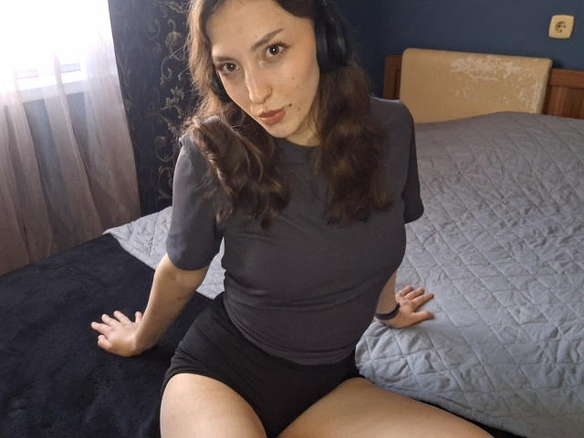 LeonieRahim, with wavy brown hair, wearing headphones, a gray t-shirt, and black shorts, sits on a bed, arms behind her, looking at the camera, showing her ass.