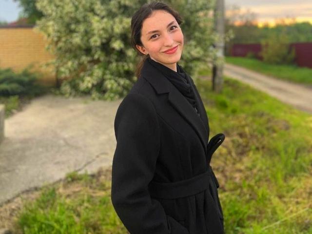 LeonieRahim, with dark hair and red lipstick, wears a black coat, standing outdoors, hands in pockets, smiling slightly, green grass, and a tree in the background.