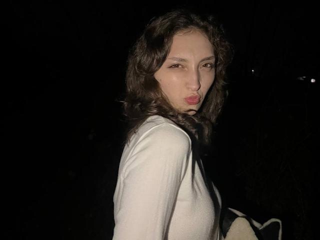 LeonieRahim has wavy brown hair, light skin, and is puckering her lips in a kissy face, wearing a white long-sleeve top, in a dark night setting.